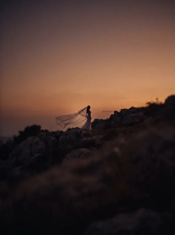 A sheer veil lifted by the wind against a dusk sky, the bride small and far away on a hilltop.